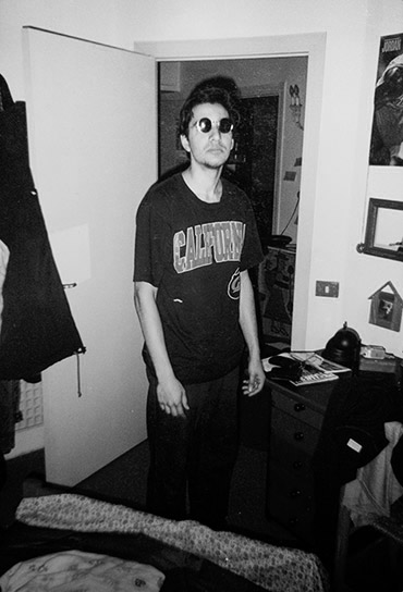A photograph showing FRANKIE wearing his trademark REVERSE SMILE in JIMMY'S place, wearing sunglasses at night. He's also wearing a dark blue shirt that says CALIFORNIA in BRIGHT YELLOW, but you can't see that because the photo is BLACK AND WHITE.