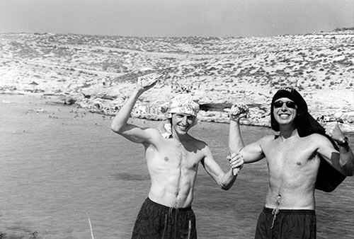 A black and white picture of Deco and Jimmy without shirts on, holding rocks, at midday in a sunny day, with a shallow bay in the background.