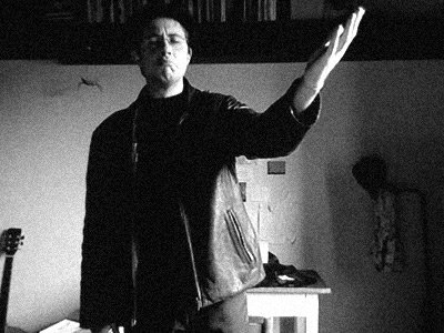 A photograph of Jimmy Boss in a room striking his famous pose with one arm up in a dismissive swing to the heavens with an Italian 'reverse smile'. There's a guitar in one corner. The picture is almost perfectly composed. A photograph of Jimmy Boss in a room striking his famous pose with one arm up in a dismissive swing to the heavens with an Italian 'reverse smile'. There's a guitar in one corner. The picture is almost perfectly composed.